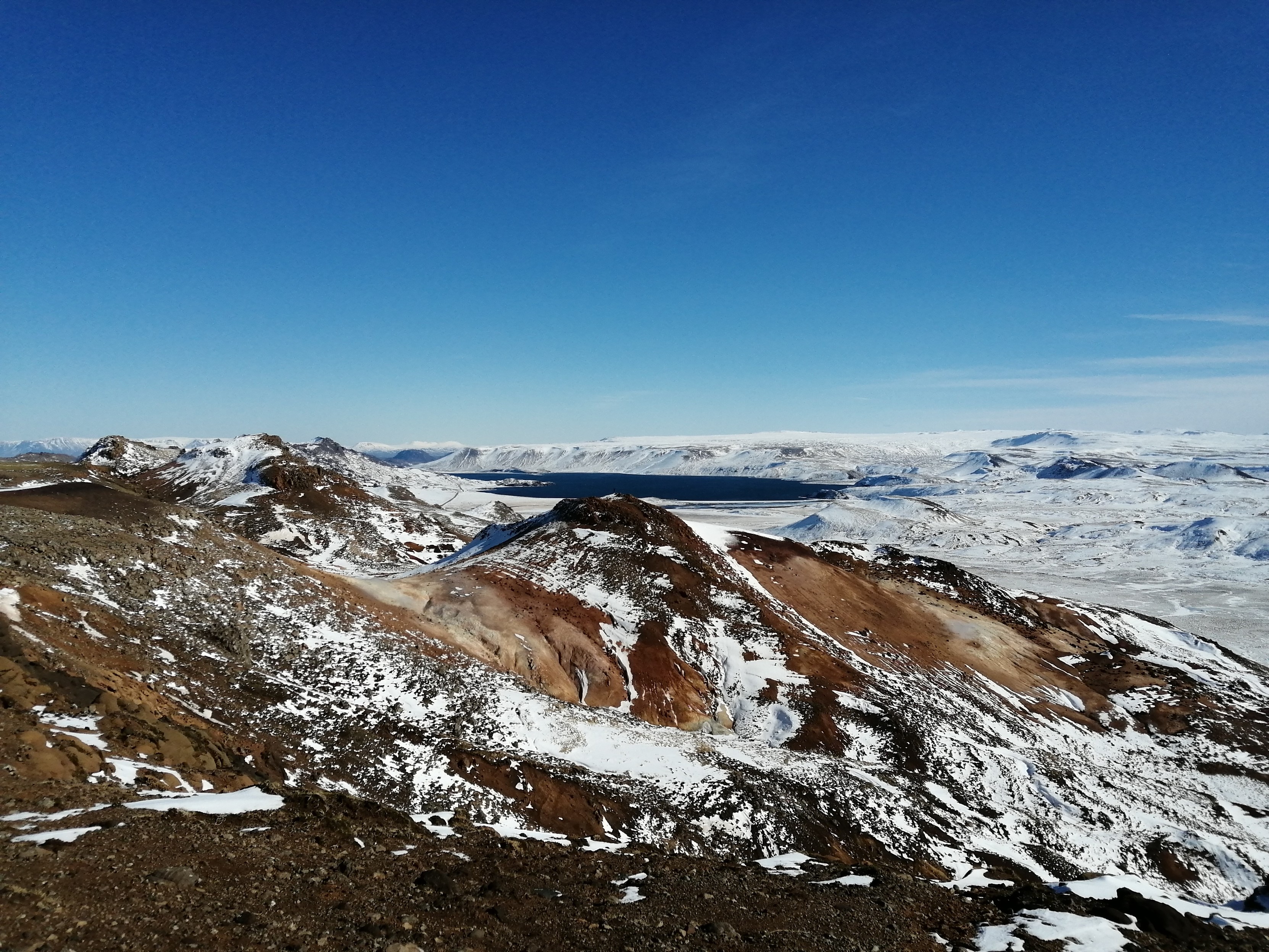 Vue générale sur les alentours du système volcanique de Krysuvik avec des montagnes peu enneigées aux teintes ocres. Ces zones regorgent de sources thermales. Tout au fond,un immense lac d'un bleu profond. Le ciel est totalement dégagé.